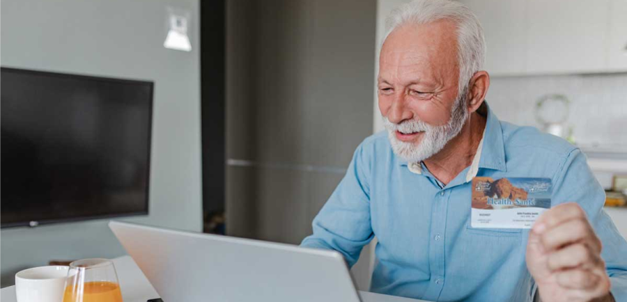 Older person holding a health insurance card while using a laptop at a table.
