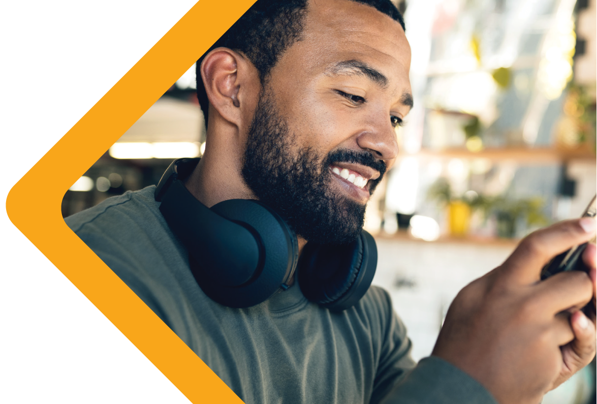 A man with a beard and headphones around his neck smiles while holding a smartphone, set against a blurred indoor background with shelves and decor, framed by an orange triangular border.
