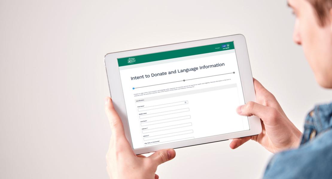 A close-up shot shows a person holding a white tablet horizontally, looking at a screen displaying a digital form titled "Intent to Donate and Language Information." The form includes fields for identification like first name, middle initial, last name, address, and city/town/community.