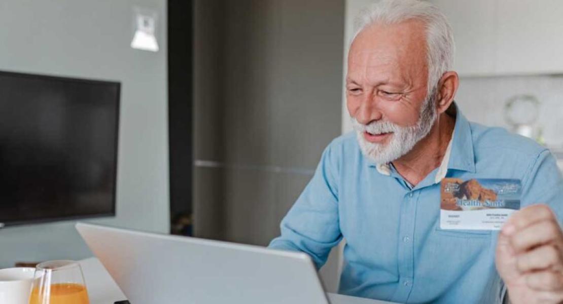 Older person holding a health card while using a laptop at a table.