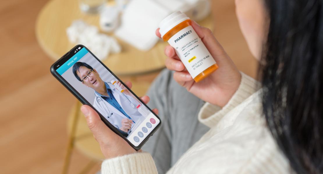 Une vue par-dessus l'épaule d'une personne utilisant un smartphone pour un rendez-vous médical virtuel tout en tenant un flacon de médicaments sur ordonnance.